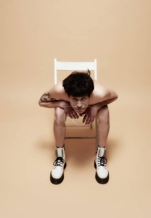 Photo for Young asian guy in underwear and stylish boots, posing shirtless on chair against light brown studio background. Concept of male body aesthetics, style, fashion, health, mens beauty - Royalty Free Image