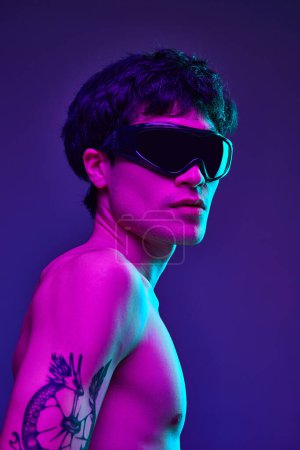 Téléchargez les photos : Portrait de jeune homme beau avec tatouage, posant torse nu dans des lunettes noires à la mode sur fond de studio violet sous la lumière du néon. Esthétique du corps masculin, style, mode, santé, concept de beauté masculine - en image libre de droit