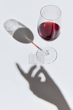 Photo for Glass filled with delicious red wine, standing against white background with female hand shadow. Celebration. Concept of taste, alcohol, wine degustation, variety, winemaking. Flat lay - Royalty Free Image