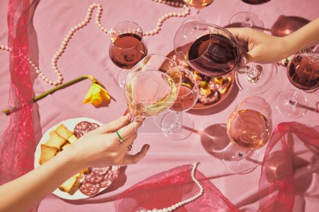 Foto de Manos femeninas tintineo vasos llenos de vino rosa sobre fondo texturizado rosa, mantel con aperitivos. Concepto de sabor, alcohol, degustación de vino, celebración, vinificación. Puesta plana - Imagen libre de derechos