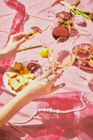 Photo for Vertical image of female hand holding glass filled with rose wine on pink textured background, tablecloth with appetizers. Taste, alcohol, wine degustation, celebration, winemaking concept. Flat lay - Royalty Free Image