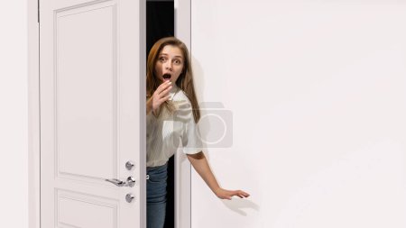 Foto de Mujer joven asomándose por la puerta gris abierta, cubriendo la boca con la mano. Pareces sorprendido, sorprendido, asombrado. Situación inesperada. Concepto de emociones, expresión facial, estilo de vida, noticias - Imagen libre de derechos