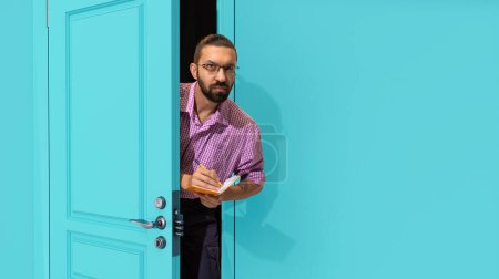 Photo for Bearded man in glasses peeking out blue door, attentively looking and making notes. Inspector, accountant, journalist. Concept of emotions, facial expression, lifestyle, employment, labour - Royalty Free Image