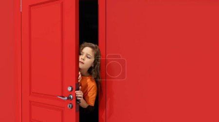 Photo for Portrait of young little girl, kid peeking out red door and attentively looking, overhearing. Playful and curious mood. Concept of childhood, emotions, facial expression, lifestyle - Royalty Free Image
