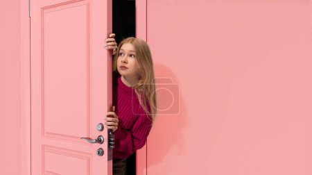 Téléchargez les photos : Belle jeune adolescente blonde regardant la porte rose et regardant avec attention, entendant des secrets. Curiosité. Concept d'émotions, expression faciale, mode de vie, jeunesse - en image libre de droit