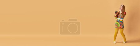 Téléchargez les photos : Portrait de jeune femme étonnante dans des vêtements élégants posant avec un appareil photo vintage sur fond orange. Style rétro, mode, beauté, élégance, concept des années 60. Style pin-up. Bannière. Espace de copie pour la publicité - en image libre de droit