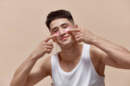 Photo for Portrait of handsome young asian guy taking care after skin, applying under eye patches against light brown studio background. Concept of male beauty, skincare, cosmetology, mens health - Royalty Free Image