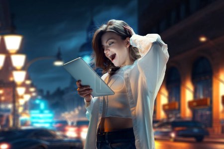 Photo for Happy and excited young girl looking on tablet with smile and positive shock, standing on street at night. Online shopping. Blurred city landscape. Modern technologies, youth, communication, business - Royalty Free Image