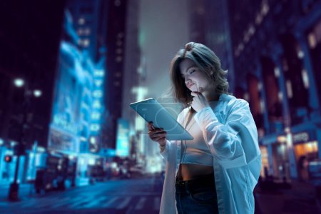 Photo for Young pretty girl standing on street at night and looking on tablet with doubtful, thoughtful face. Blurred city background. Online shopping. Modern technologies, youth, communication and business - Royalty Free Image