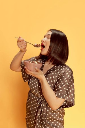 Photo for Portrait of beautiful, young, asian girl in stylish silk pajamas, eating cereal with milk against yellow studio background. Delicious breakfast. Concept of emotions, lifestyle, youth, comfort - Royalty Free Image