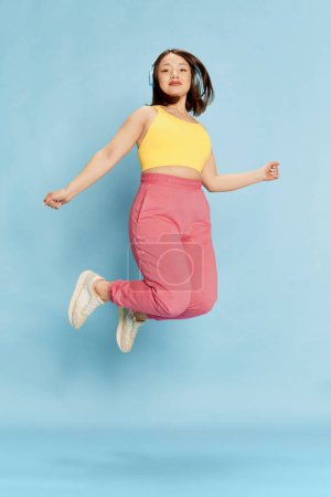 Foto de Retrato de larga duración de una joven asiática en top amarillo y pantalón rosa, escuchando música en auriculares y saltando sobre fondo azul del estudio. Concepto de emociones, estilo de vida, juventud - Imagen libre de derechos
