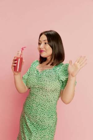 Portrait of positive, happy, young, asian girl in stylish green dress, drinking berry smoothie against pink studio background. Delicious food. Concept of emotions, lifestyle, youth, diet, health
