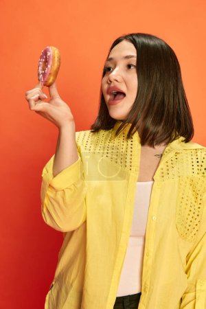 Foto de Amante de los postres. Retrato de joven, bonita, chica asiática en blusa amarilla, sosteniendo rosquilla rosa, posando sobre fondo de estudio naranja. Emoción. Concepto de emociones, estilo de vida, juventud, moda - Imagen libre de derechos