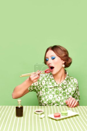Téléchargez les photos : Jolie, élégante, jeune fille mangeant des macarons sucrés avec des baguettes sur fond de studio vert. Goût de sauce soja. Photographie pop art culinaire. Concept de style rétro, vision créative, imagination. - en image libre de droit