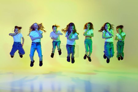 Téléchargez les photos : Performance. Petites filles, enfants en tenue sportive décontractée dansant, sautant sur fond de studio vert sous la lumière du néon. Concept d'enfance, passe-temps, mode de vie sportif, action et mouvement - en image libre de droit