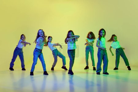 Foto de Imagen dinámica de niñas, niños bailando sobre fondo de estudio verde con luz de neón. Ropa casual. Estilo hip-hop. Concepto de infancia, afición, estilo de vida deportivo, acción y movimiento - Imagen libre de derechos