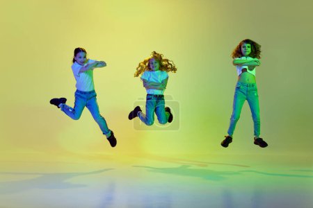 Photo for Three active, little girls, children jumping, dancing in casual clothes against green studio background in neon light. Concept of childhood, hobby, sportive lifestyle, fashion, action and motion - Royalty Free Image