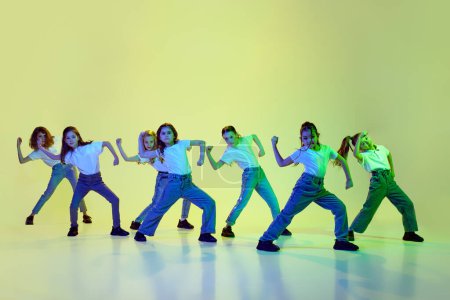 Photo for Artistic, talented little girls, children in casual sporty clothes dancing hip-hop against green studio background in neon light. Concept of childhood, hobby, sportive lifestyle, friendship, education - Royalty Free Image