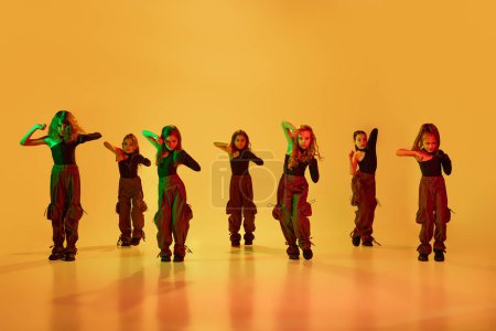 Téléchargez les photos : Groupe de petites danseuses, filles, enfants en vêtements de sport élégants dansant sur fond de studio jaune sous la lumière du néon. Concept d'enfance, passe-temps, mode de vie sportive, mode, éducation - en image libre de droit