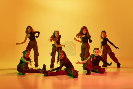 Photo for Art. Group of children, little girls in sportive casual style clothes dancing hip-hop, contemp dance against yellow studio background in neon light. Concept of childhood, hobby, sportive lifestyle - Royalty Free Image