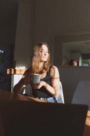 Téléchargez les photos : Portrait de jeune belle femme passant du temps confortable à la maison le jour avec la lumière du soleil. Assis à table et buvant le café du matin. Concept de vie à la maison, émotions, vacances, week-ends - en image libre de droit