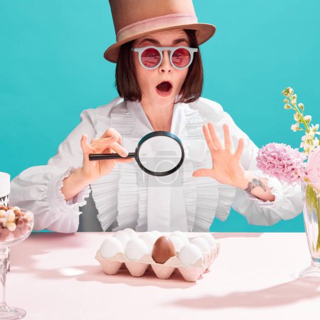 Photo for Shocked woman in sunglasses and cylinder hat emotionally looking in magnifying glass at chocolate eggs among ordinary. Concept of pop art, creativity, food, inspiration, holidays, emotions - Royalty Free Image