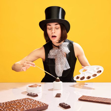 Foto de Mujer, en imagen de pintor de alimentos, en sombrero de cilindro haciendo dulces de chocolate, pintando caramelos sobre fondo amarillo. Obra maestra de la confitería. Concepto de arte pop, creatividad, comida, inspiración - Imagen libre de derechos