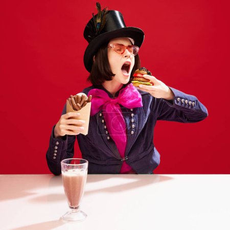 Téléchargez les photos : Femme élégante en chapeau de cylindre et tailcoat avec arc rose manger burger et bâtons de chocolat sur fond rouge. Doux et salé. Concept de pop art, créativité, gastronomie, inspiration - en image libre de droit