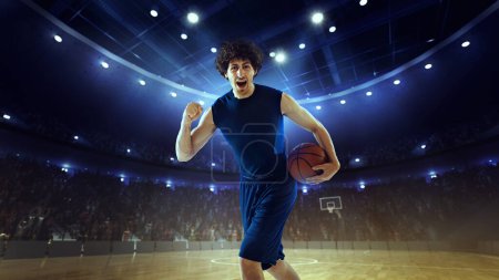 Photo for Win, success. Young man, basketball player in uniform standing with ball on 3D stadium under spotlights with blurred audience. Concept of professional sport, competition, action and motion, game - Royalty Free Image
