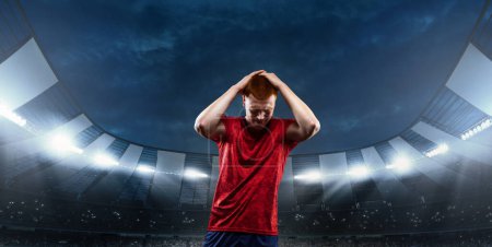 Photo for Loosing game. Young man, professional sportsman in red uniform holding head with hands in despair and sadness at 3D stadium with flashlights. Concept of sport, competition, game, emotions - Royalty Free Image