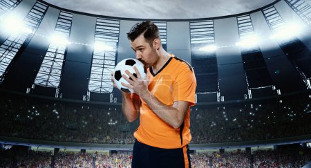 Photo for Man, professional football sportsman in orange uniform kissing ball for successful winning game, standing on 3D field with spotlights. Concept of sport, competition, game, emotions - Royalty Free Image