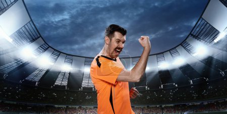 Téléchargez les photos : Homme émotionnel, sportif professionnel de football en uniforme orange debout avec regard gagnant sur le terrain 3D avec projecteurs. Concept de sport, compétition, jeu, émotions, succès - en image libre de droit