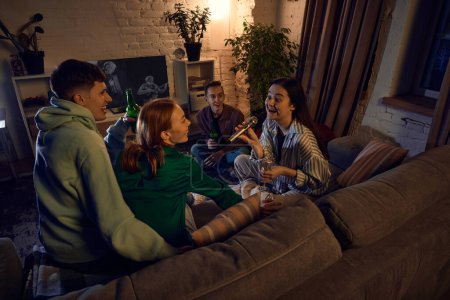 Foto de Grupo de amigos felices y sonrientes sentados en el sofá en casa por la noche y cantando karaoke, hablando, bebiendo cerveza y divirtiéndose. Concepto de amistad, ocio, fines de semana, diversión, emociones - Imagen libre de derechos
