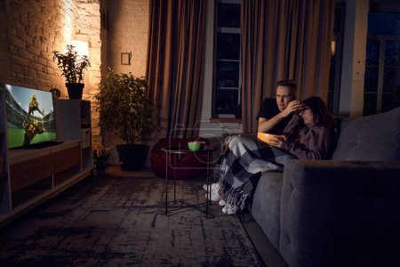 Téléchargez les photos : Jeune couple, homme et femme passent du temps ensemble à la maison le soir, assis sur le canapé et regardant un match de football en ligne. Je perds la partie. Concept d'amitié, loisirs, week-ends, émotions - en image libre de droit