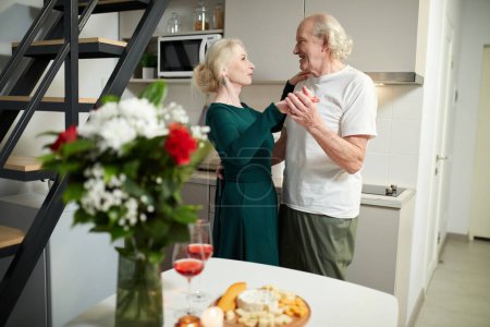 Photo for Beautiful, happy senior couple, man and woman celebrating marriage anniversary, having romantic dinner in kitchen at home, dancing. Concept of love, relationship, family, emotions, support and care - Royalty Free Image
