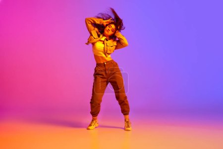 Foto de Chica joven flexible, artística, bailarina profesional en ropa deportiva bailando hip-hop contra el degradado fondo rosa púrpura en luz de neón. Concepto de danza contemporánea, juventud, hobby, acción, movimiento - Imagen libre de derechos