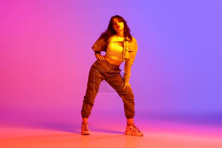 Téléchargez les photos : Artistique, belle, jeune fille dansant hip-hop sur fond violet rose dégradé dans la lumière au néon. Modèle en vêtements décontractés. Concept de danse contemporaine, jeunesse, passe-temps, action et mouvement - en image libre de droit