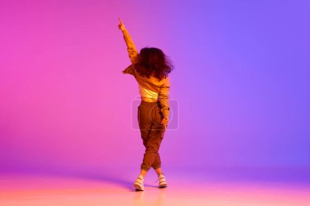 Foto de Dale la vuelta. Chica joven., bailarina profesional en ropa de estilo deportivo que actúa contra el gradiente rosa púrpura fondo en luz de neón. Concepto de danza contemporánea, juventud, hobby, acción y movimiento - Imagen libre de derechos