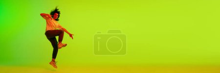 Foto de Chica joven en ropa deportiva bailando hip-hop tagainst gradiente verde amarillo fondo en luz de neón. Concepto de danza contemporánea, juventud, hobby, acción y movimiento. Banner. Copiar espacio para anuncio - Imagen libre de derechos