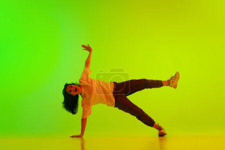Foto de Chica joven artística, flexible, talentosa, bailarina de hip-hop que actúa contra el fondo verde degradado amarillo en luz de neón. Concepto de danza contemporánea, juventud, hobby, acción y movimiento - Imagen libre de derechos
