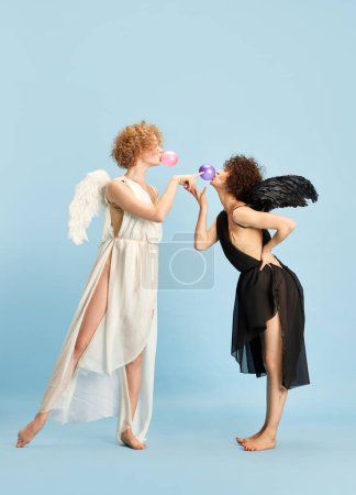 Téléchargez les photos : Tandem de mal et de bonté. Portrait de deux belles jeunes femmes en images d'ange et de démon sur fond bleu. Concept d'émotions, comparaison, créativité, beauté, valeurs humaines - en image libre de droit