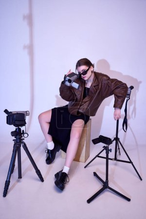 Photo for Filming industry. Stylish young girl in leather jacket and trendy sunglasses posing with retro video camera. Concept of fashion, style, retro and vintage, gadgets, beauty, technology, 80s, 90s - Royalty Free Image