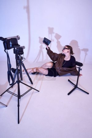 Foto de Elegante chica joven en chaqueta de cuero, vestido y gafas de sol de moda posando con cámaras de vídeo retro, filmación, haciendo contenido. Concepto de moda, 80s, 90s style, retro y vintage, gadgets, beauty - Imagen libre de derechos
