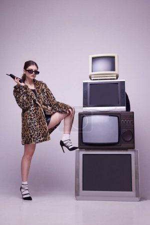 Photo for Television creative manager. Young girl in stylish animal print coat and sunglasses posing near retro TV sets against grey background. Concept of fashion, 80s, 90s style, retro and vintage, mass media - Royalty Free Image