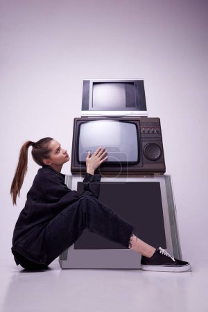 Téléchargez les photos : Addiction aux médias de masse. Jolie jeune fille en vêtements noirs assise près des téléviseurs rétro sur fond de studio gris. Mode, années 80, 90 style, rétro et vintage, gadgets, beauté, concept technologique - en image libre de droit