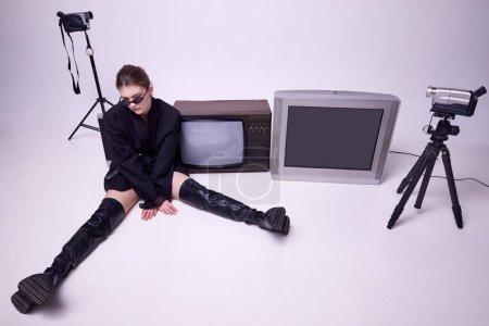 Photo for Young girl in stylish black jacket, leather boots and sunglasses sitting on floor around retro TV sets and camera. Concept of fashion, 80s, 90s style, retro and vintage, gadgets, mass media - Royalty Free Image