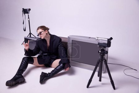 Téléchargez les photos : Jeune fille en veste noire élégante, bottes en cuir et lunettes de soleil assis sur le sol autour des téléviseurs rétro et de la caméra. Concept de mode, années 80, 90 style, rétro et vintage, gadgets, beauté, technologie - en image libre de droit