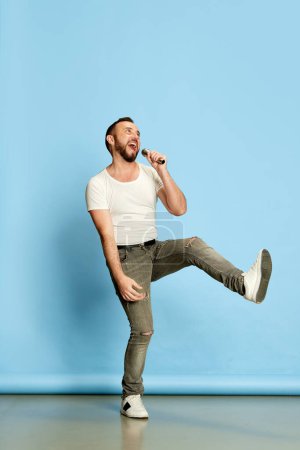 Photo for Full-length portrait of bearded happy man in casual white t-shirt singing in microphone, having fun against blue studio background. Concept of human emotions, lifestyle, facial expression. Ad - Royalty Free Image