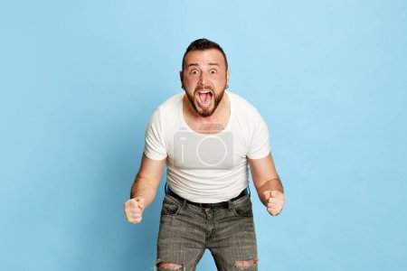 Téléchargez les photos : Portrait d'homme barbu en t-shirt blanc posant avec des émotions excitées, positives et gagnantes sur fond de studio bleu. Concept d'émotions humaines, mode de vie, expression faciale, succès. Publicité - en image libre de droit