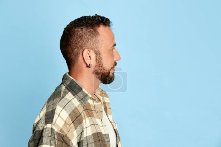 Téléchargez les photos : Portrait latéral de l'homme barbu en chemise à carreaux regardant loin, posant sur fond de studio bleu. Concept d'émotions humaines, style de vie, expression faciale. Publicité - en image libre de droit